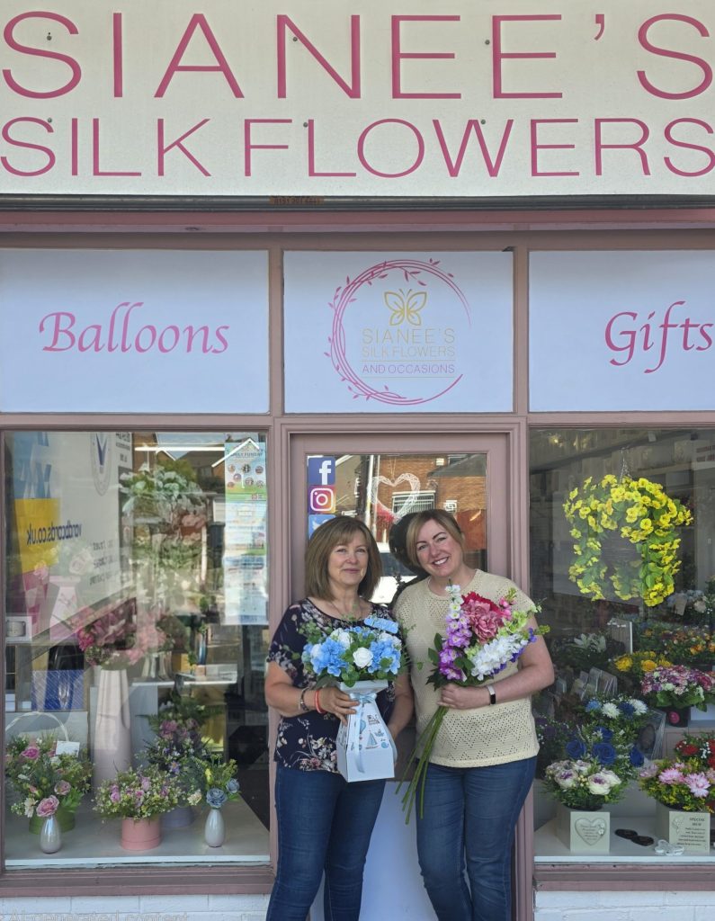A storefront with the signs "SIANEE'S SILK FLOWERS", "Balloons", and "Gifts" is shown. Two women are standing in the doorway, each holding a bouquet of artificial flowers. One woman on the left holds a bouquet of blue an white silk flowers, whilst the other on the right holds a colourful bouquet of pink, purple and cream silk flowers. The shop's windows are filled with various silk flower arrangements.
