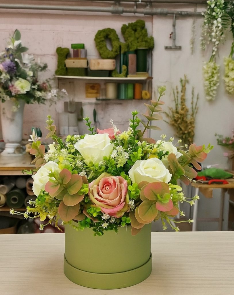 A silk flower arrangement is displayed in a round, light green box. The silk flower arrangement features a mix of white and pale pink artificial roses, surrounded by various green and red foliage. There are also smaller white and yellow filler faux flowers. The background is a floral studio with shelves holding other floral items.